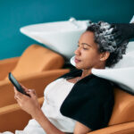 African American woman using smart phone while getting her hair washed at hair salon.